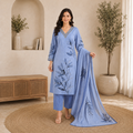 Woman in a blue embroidered outfit standing in a room with decorative plants and furniture.